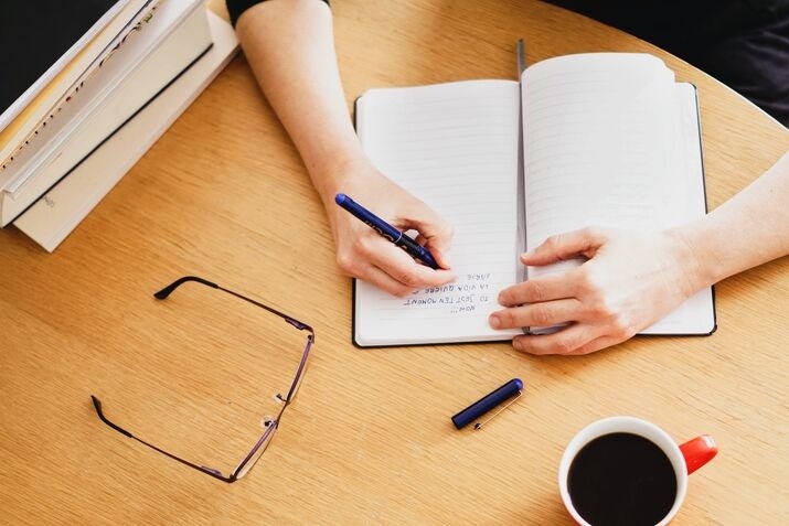 Person writing notes in a notebook beside books, glasses, and a coffee cup.