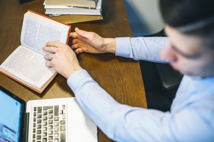 Person proofreading a book manuscript while working on a laptop at a desk