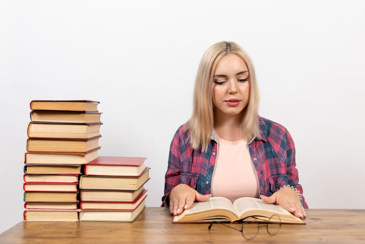 Woman reading a book with a stack of books beside her during proofreading
